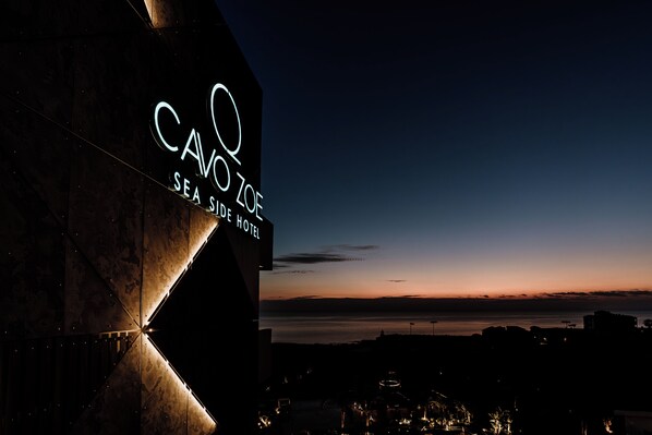 Un letrero de hotel con el nombre "CAVO ZOE SEA SIDE HOTEL" contra un cielo al anochecer.