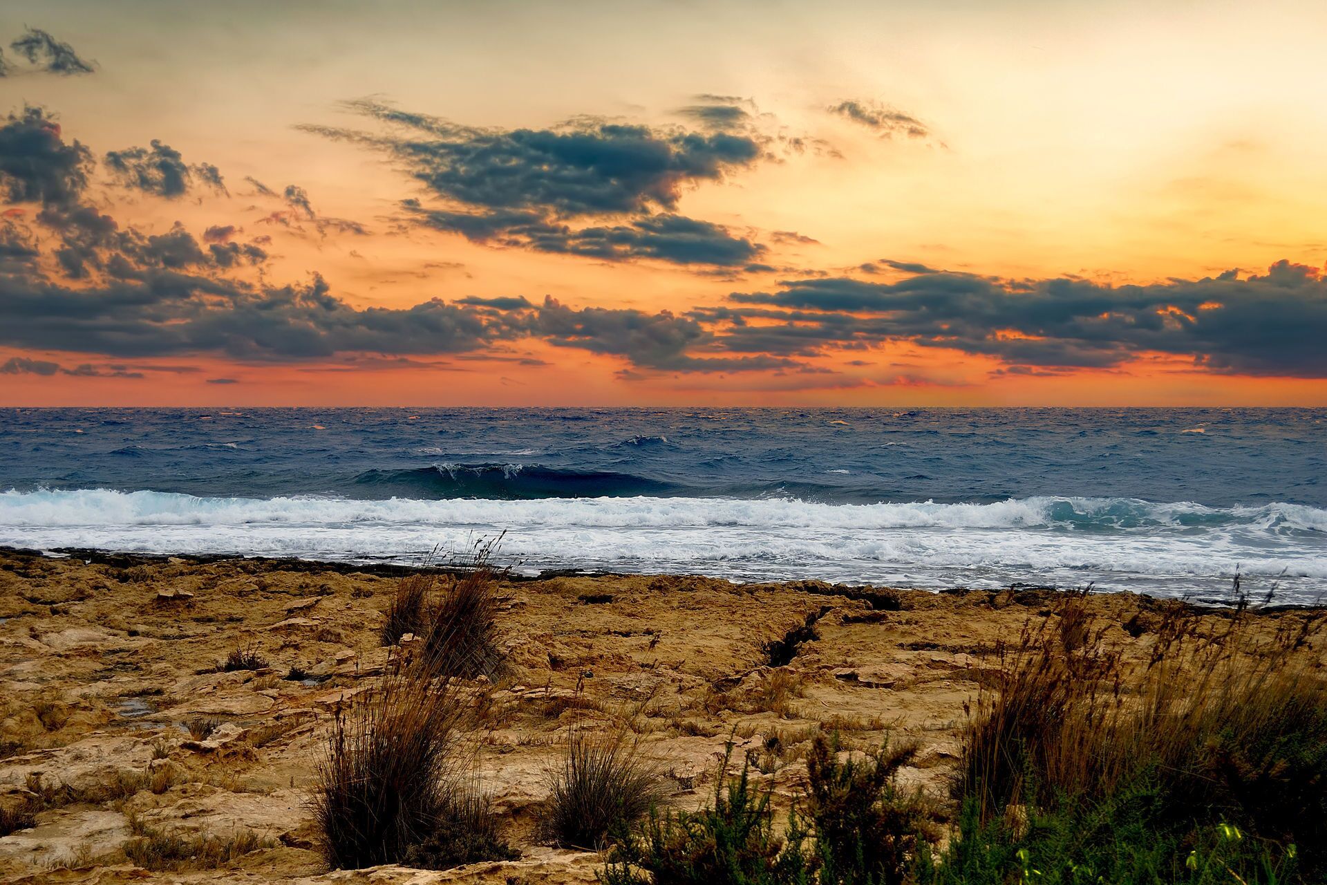 A beach with waves at sunset, rocky terrain, and sparse vegetation.