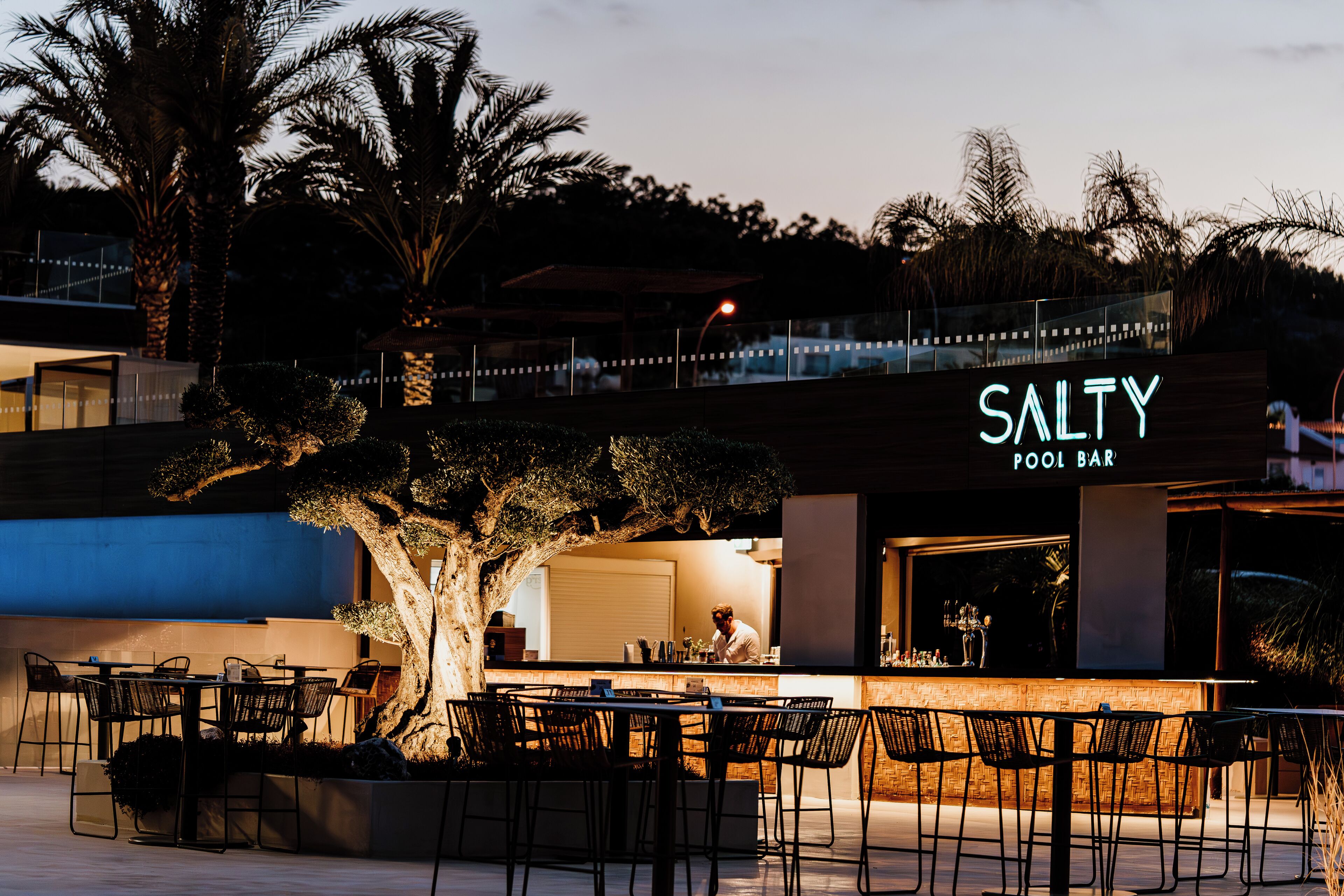 Un bar situé au bord de la piscine, appelé SALTY, avec des places assises en plein air et un grand arbre au premier plan.