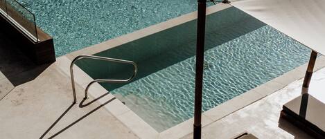 Une piscine à l’eau claire, une échelle en métal et un espace ombragé avec des chaises longues.