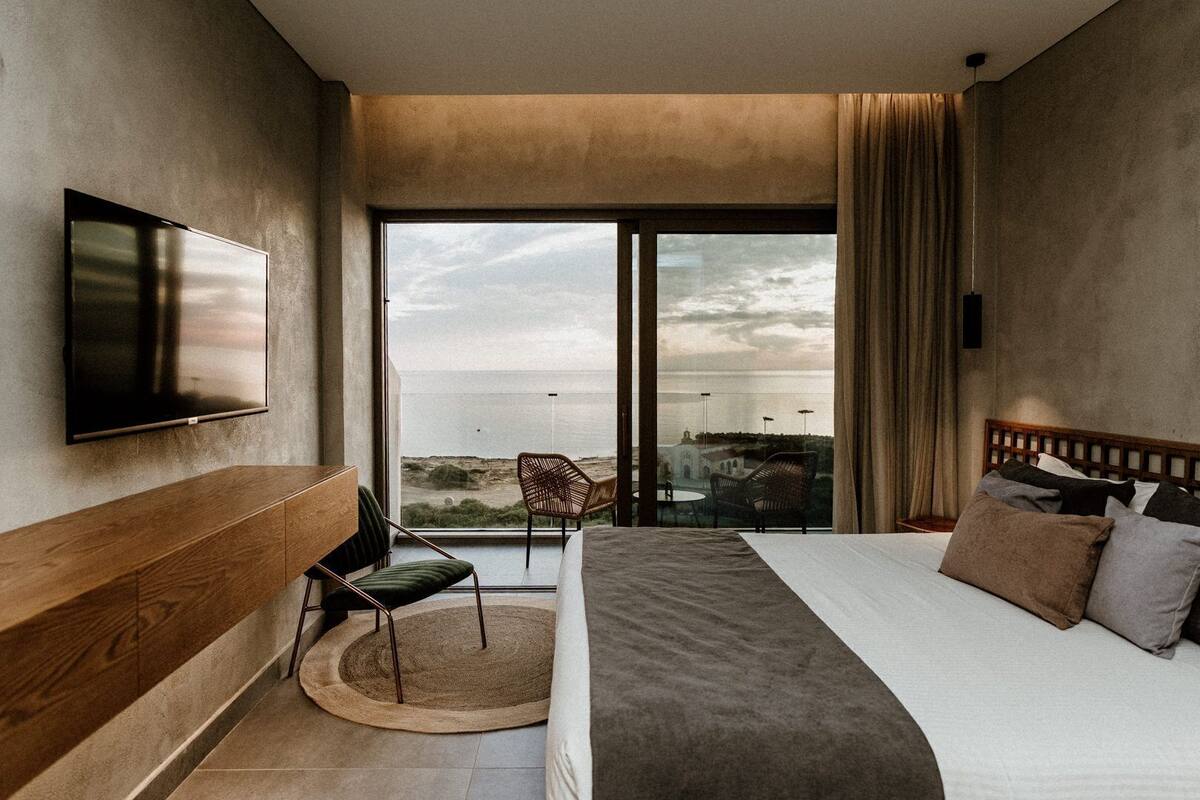 Ein modernes Hotelzimmer mit einem großen Bett, einem an der Wand befestigten Fernseher, einem runden Teppich und Meerblick durch große Fenster.