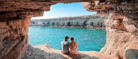 Una pareja sentada en un risco con vista al mar azul y despejado, con acantilados al fondo.
