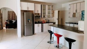 A compact kitchen with a refrigerator, microwave, and a water dispenser. There are two bar stools and a small dining table. - WestPoint Quays (Harare)