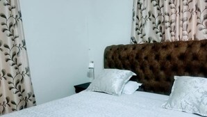 A neatly made bed with a white patterned bedspread, a tufted brown headboard, and two white pillows. - WestPoint Quays (Harare)