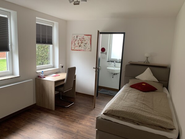 A bedroom with a bed, a desk, and a chair. There is a window with blinds, a mirror, and a sink.