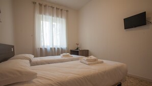Habitación de hotel con dos camas, un televisor de pantalla plana montado en la pared y una ventana con cortinas translúcidas.