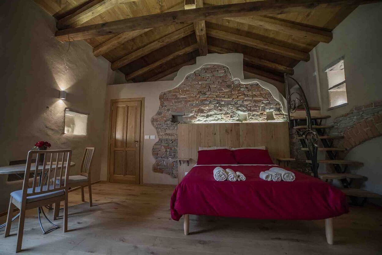 A rustic bedroom with a stone fireplace, wooden ceiling beams, a bed with red bedding, and a dining area with chairs.