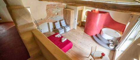 A loft bedroom with a sloped ceiling, exposed wooden beams, a brick wall, a red sofa, a small round table, and a white bathtub.