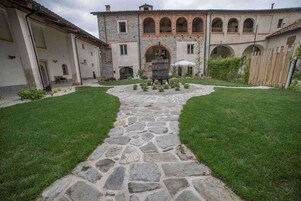 Un patio con un sendero de piedra que lleva a una fuente central, rodeado por un edificio con ventanas de arco y techo de tejas.