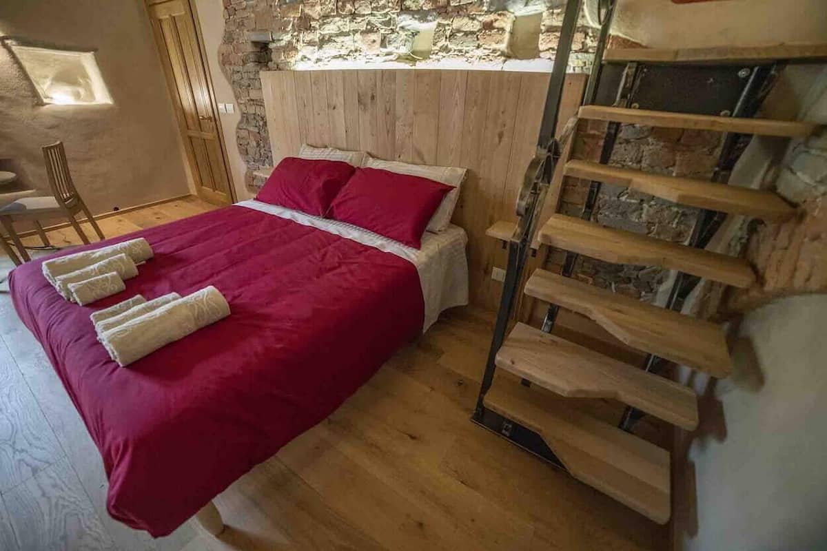 A bedroom with a wooden bed, red bedding, a wooden ladder, and a stone wall.