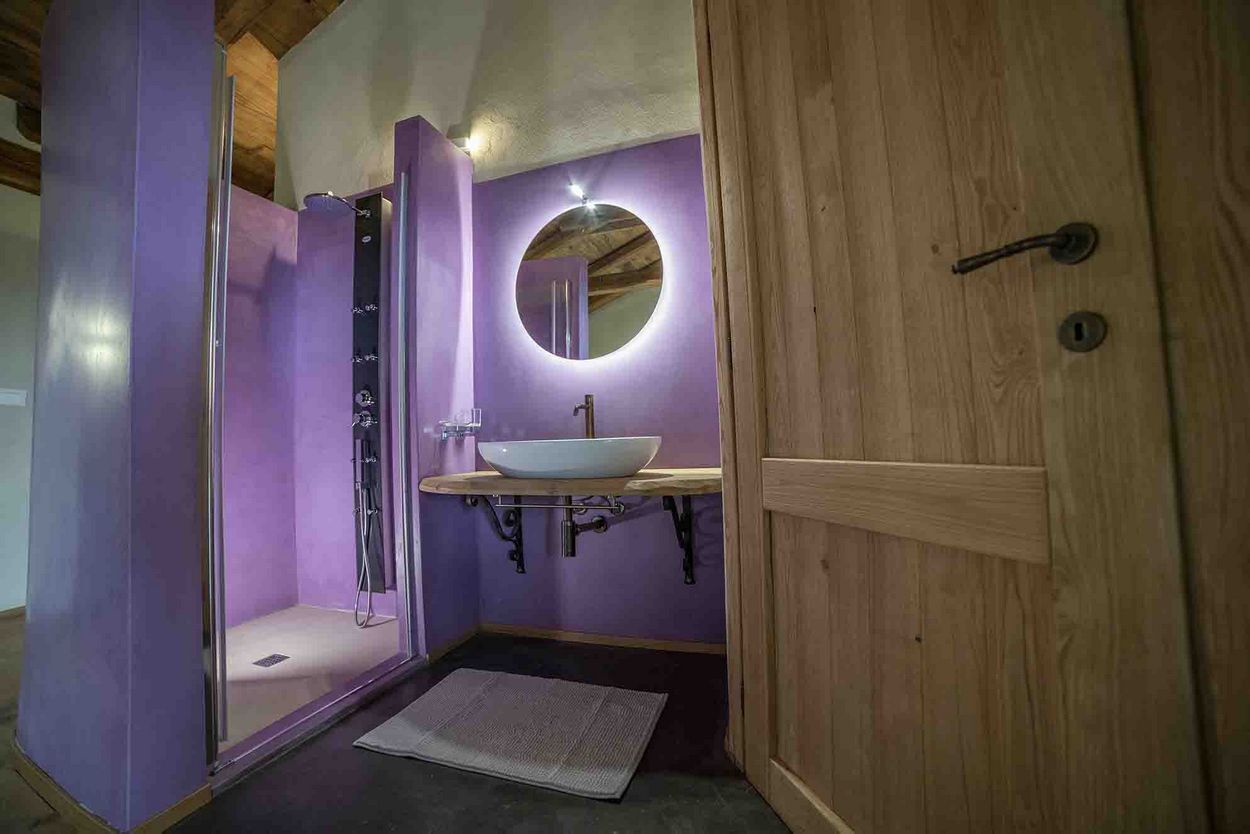 Un baño con pared morada, puerta de madera, lavabo blanco y cabina de ducha.