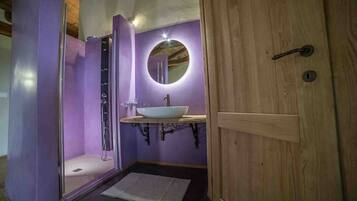 Un baño con pared morada, puerta de madera, lavabo blanco y cabina de ducha.