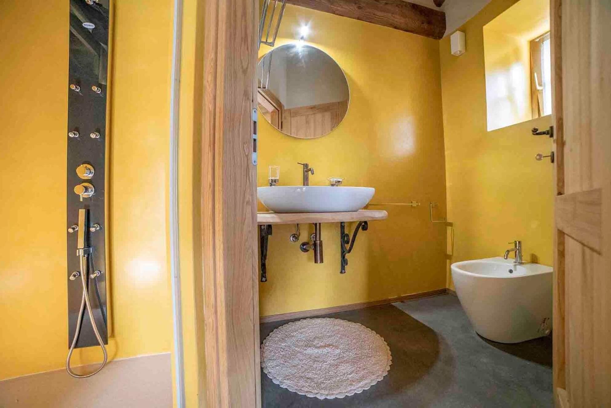 A bathroom with a yellow wall, a round mirror, a white sink, a bidet, and a wooden door frame.