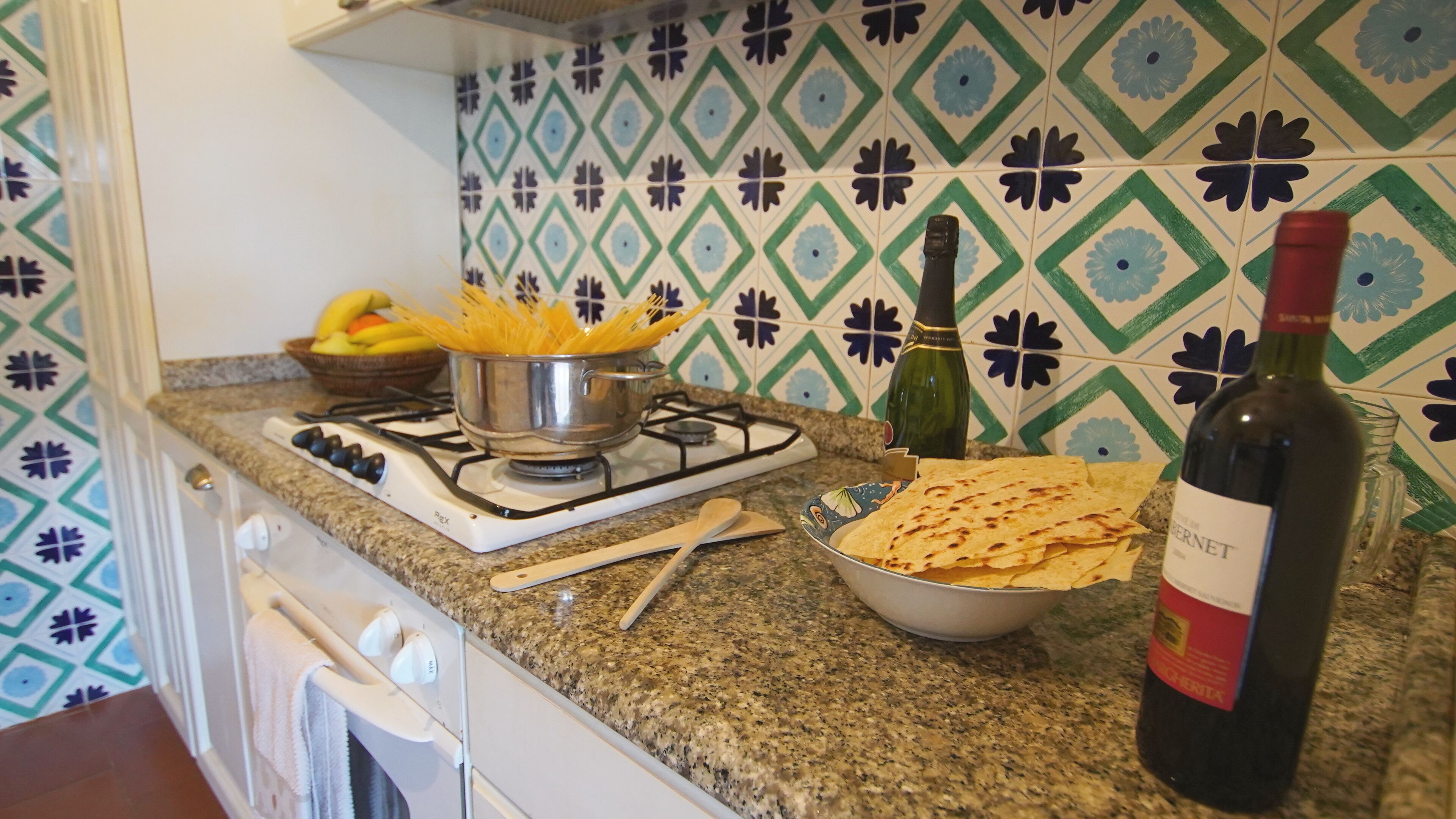 Une cuisine équipée d’une cuisinière à gaz, un plat de pâtes, une bouteille de vin et un bol de tortillas.