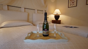 Cama com lençóis brancos, uma garrafa de vinho, duas taças e um suporte sobre um tapete branco texturizado.