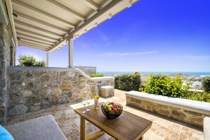 A stone wall, a wooden table with a fruit bowl and wine glasses, and a view of the sea.