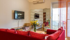 Ein Wohnzimmer mit einem roten Sofa, einem flachbildfernseher an der Wand, einer Vase mit gelben Blumen auf dem Couchtisch und einer Glastür, die auf einen Balkon führt.