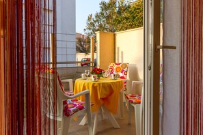 Um pátio externo com toalha de mesa amarela, cadeiras brancas e uma pequena mesa posta com um vaso de flores e taças de vinho.