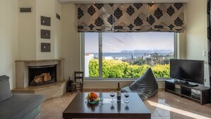 A living room with a fireplace, a television, and a large window with a view of greenery and buildings. - Villa Familia Chania (Chania)