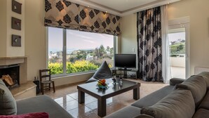 A living room with a fireplace, a television, a coffee table with fruit, and a large window with a view. - Villa Familia Chania (Chania)