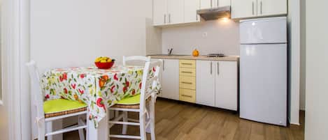 A small kitchen with white cabinets, a white refrigerator, and a table with a fruit bowl.