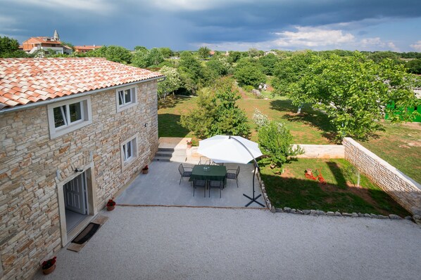 Un edificio de piedra con tejado de baldosas, un patio con mesa y sillas, y vista a un paisaje verde con árboles y un edificio distante.