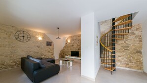A living room with a stone wall, a black sofa, a coffee table, and a wooden spiral staircase. - Stone house Furlani with a lovely garden (Svetvincenat)