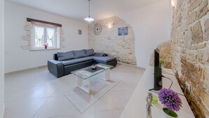 A living room with a sectional sofa, a glass coffee table, a stone wall, a clock, and a television. - Stone house Furlani with a lovely garden (Svetvincenat)