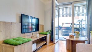 Un salon moderne avec un téléviseur à écran plat, des meubles en bois, un coussin vert, une petite table avec un vase et des biscuits, et une porte-fenêtre coulissante donnant sur l’extérieur.