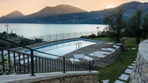 A swimming pool with a wooden deck and lounge chairs, surrounded by a stone wall and a metal railing.