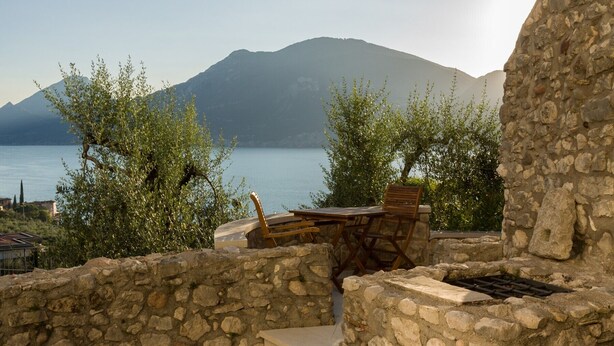 A stone-walled outdoor seating area with a table and chairs, overlooking a body of water and a mountain.