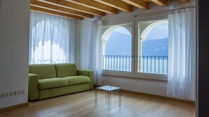 A room with a green sofa, a small table, and large windows with a view of the sea and mountains.