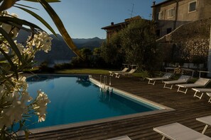 A pool with lounge chairs and a wooden deck, surrounded by greenery and a stone building.