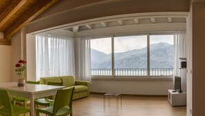 A room with a dining table, green chairs, a sofa, a small table, and a TV. It has large windows with a mountain view.