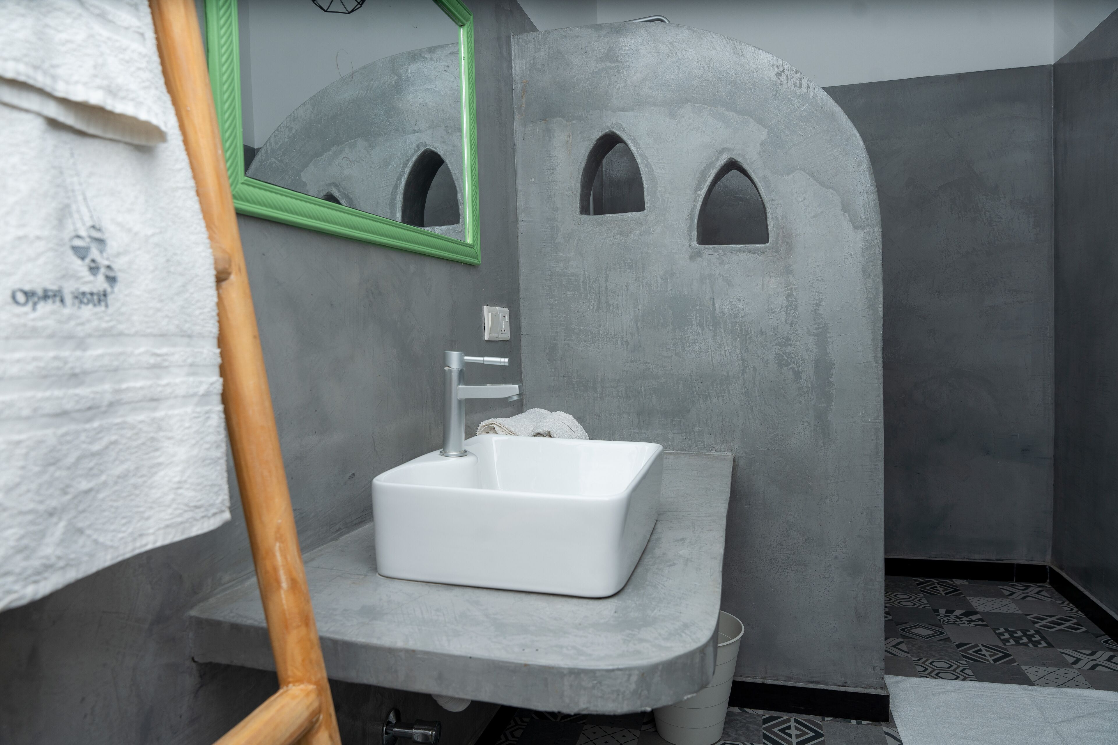 A bathroom with a white sink, a green-framed mirror, and a towel hanging on a wooden rack.