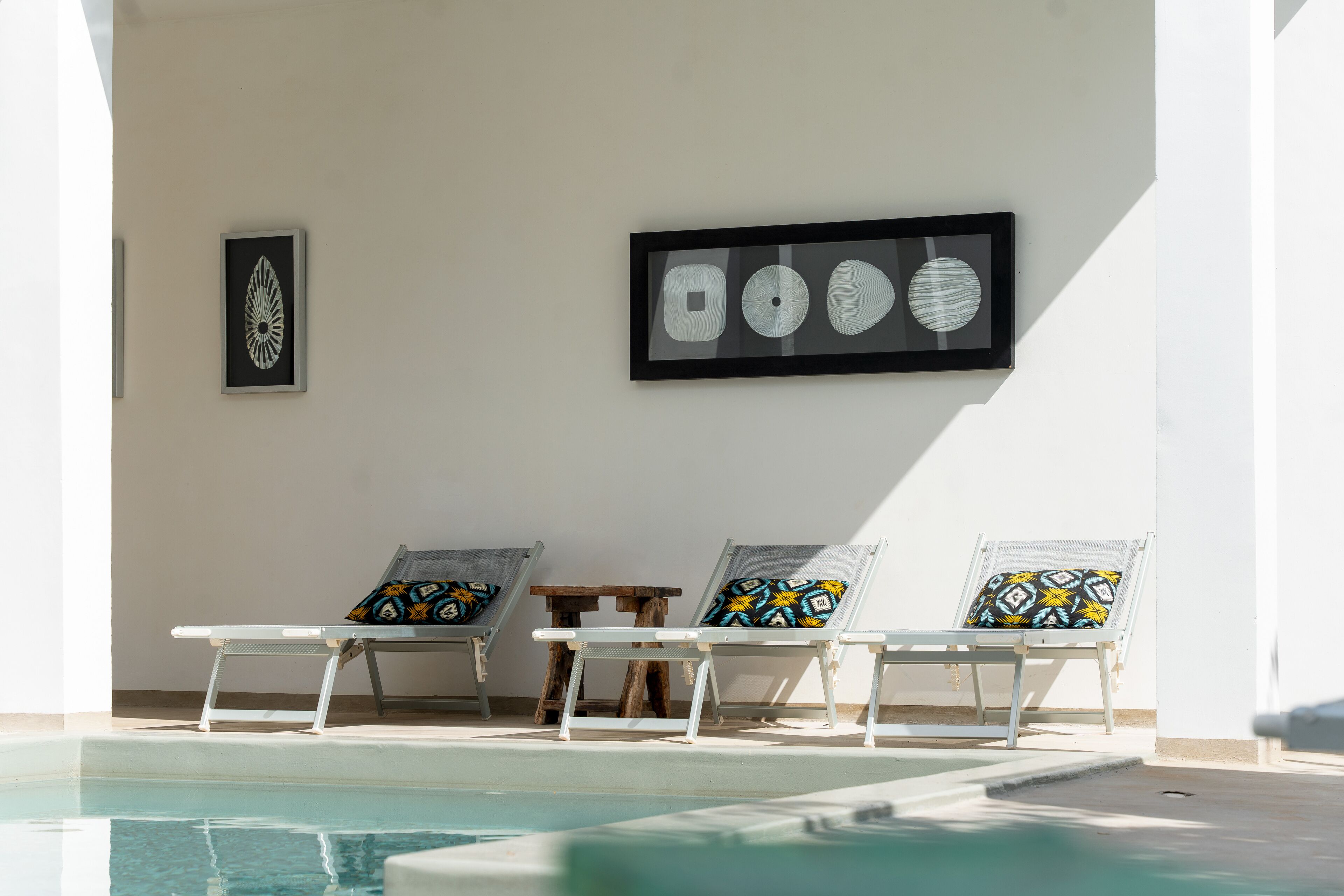 A poolside area with three sun loungers, a small wooden table, and a framed artwork on the wall.