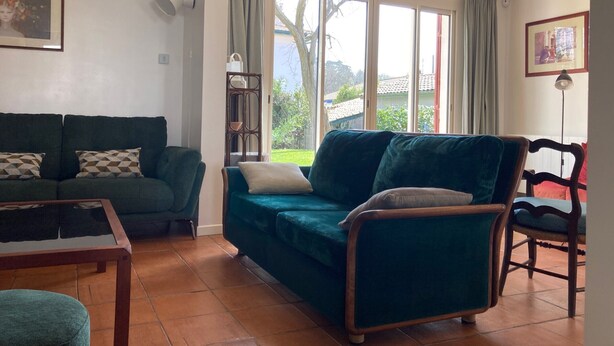 A living room with a sofa, a chair, a table, and a view of the outdoors through large windows. - Charming 3-Star Beach House, Ideal for Families, with Garden and Parking (Hendaye)
