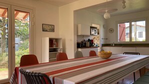 A dining area with a long table, chairs, a kitchen in the background, and a sliding glass door leading outside. - Charming 3-Star Beach House, Ideal for Families, with Garden and Parking (Hendaye)