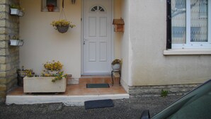 A house with a white door, a small window with a white frame, and a number '3' above the door. There are flower pots on the steps and a hanging basket with flowers.