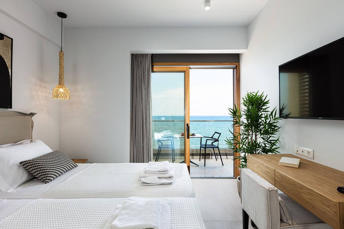 A modern hotel room with a balcony, a bed with white linens, a wooden desk, and a television on the wall.