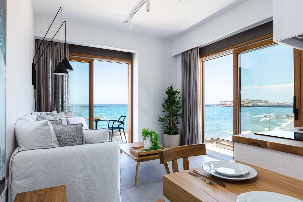 A modern living room with a sofa, dining table, and large windows overlooking the sea.