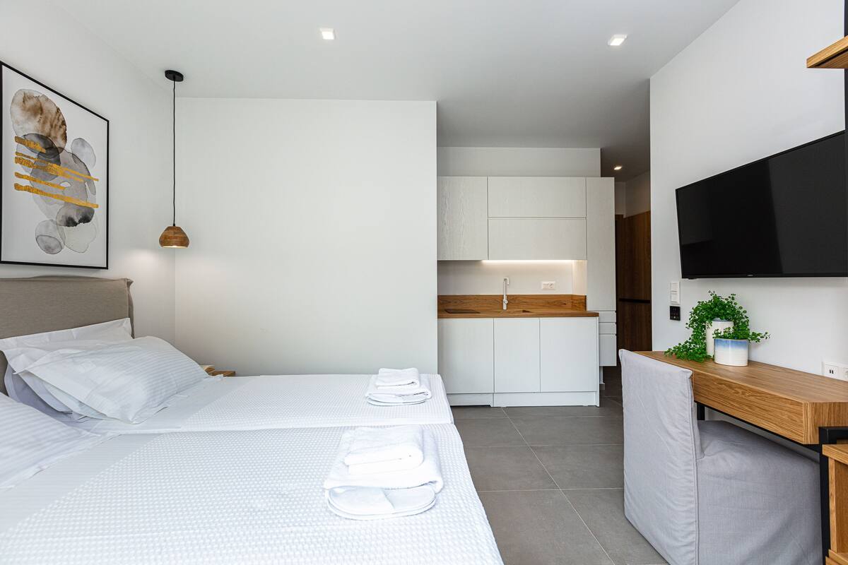 A modern hotel room with two beds, a TV, a desk, and a kitchen area.