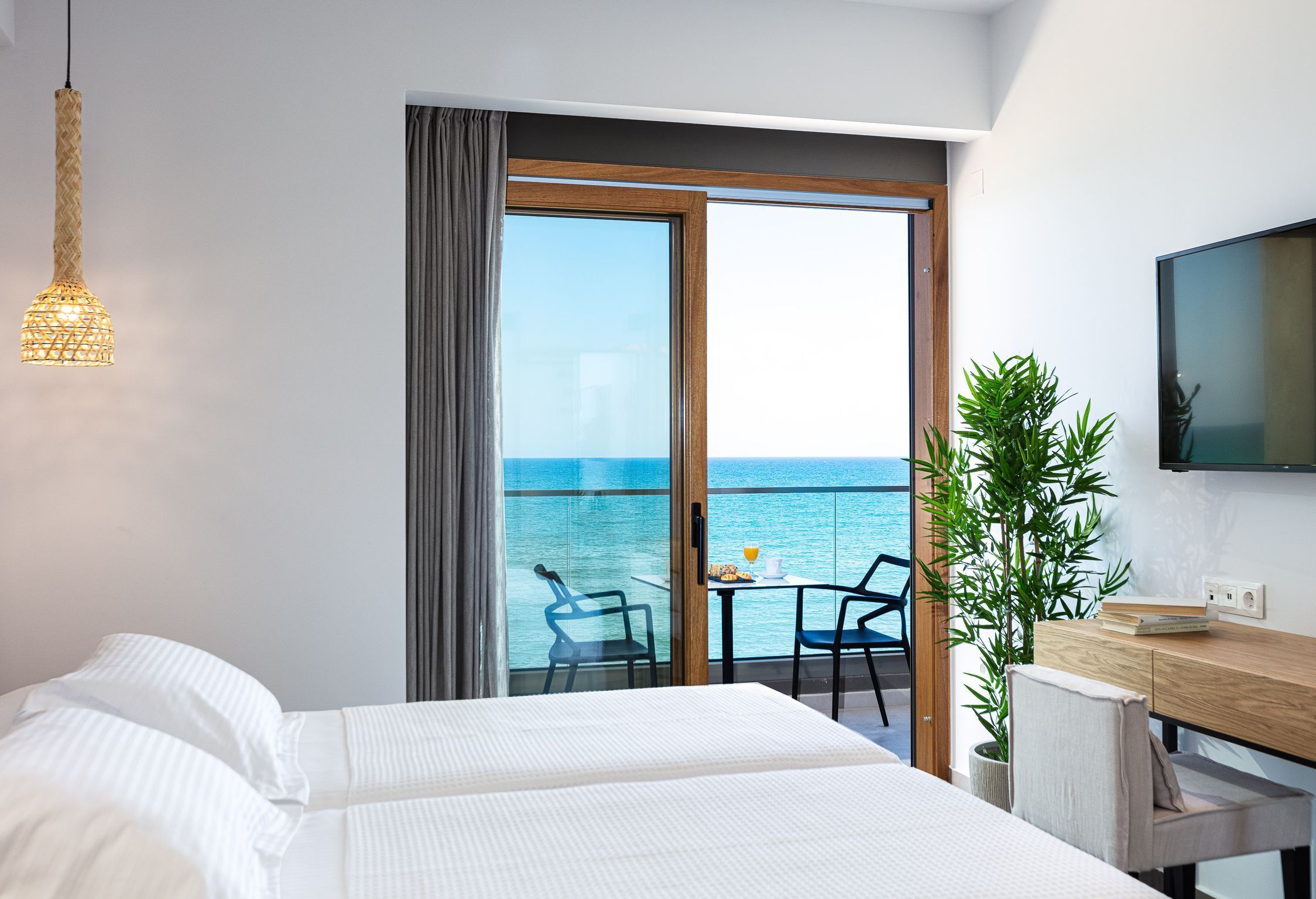 A hotel room with a bed, a desk, a chair, a TV, a plant, and a balcony with a view of the sea.