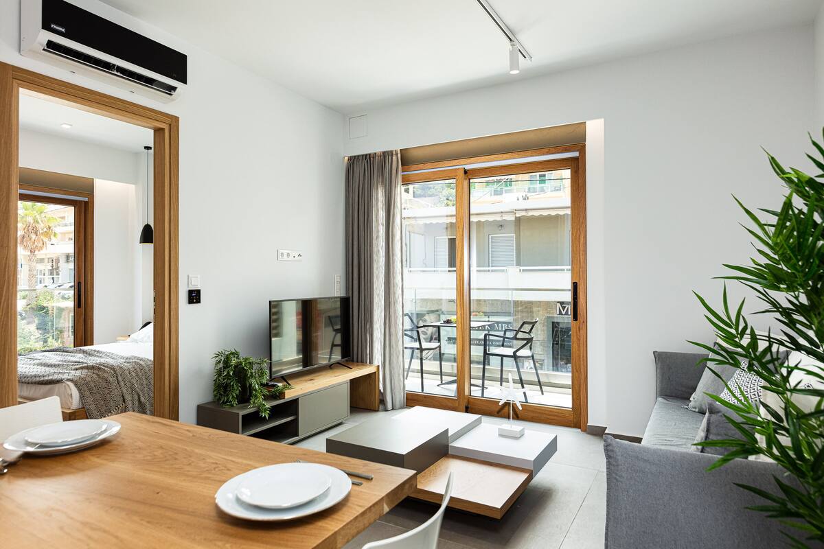 A modern hotel room with a wooden dining table, a sofa, a TV, and a balcony with a view of the city.