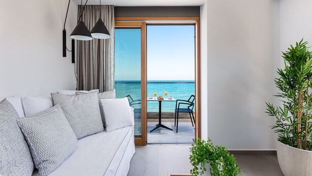 A modern living room with a white sofa, a dining table with chairs, and a view of the sea through a glass door. - North Coast Seaside Suites (Rethymno)