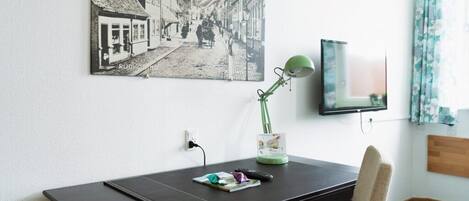 Una habitación con un escritorio, una fotografía antigua en la pared, una lámpara de escritorio verde y un televisor.