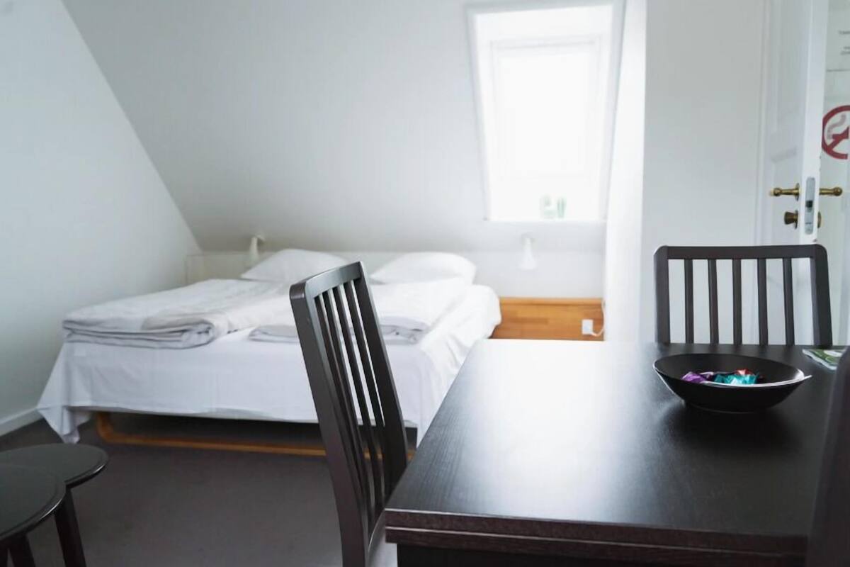 Una habitación con una cama, una mesa con un cuenco, una silla y un ventanal.
