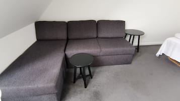A corner seating area with a grey sectional sofa, two small round tables, and a white bed in a room with grey carpet.