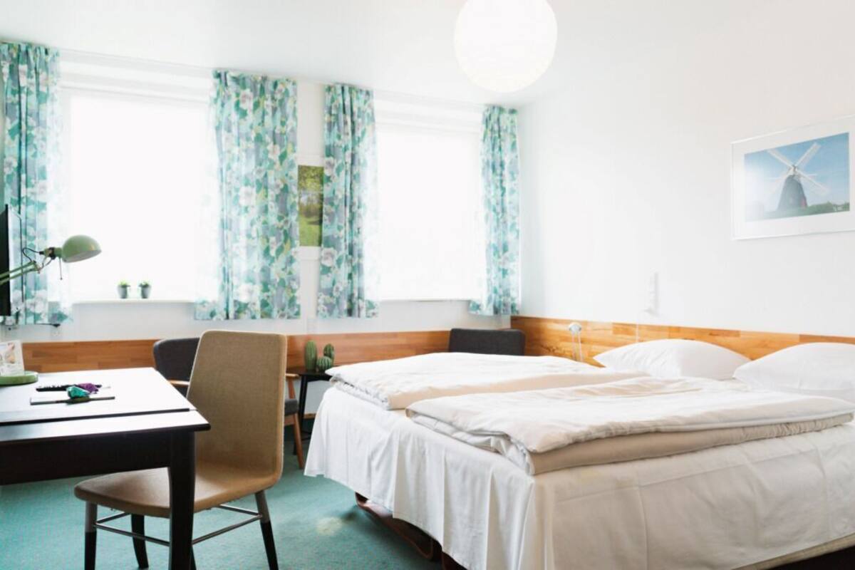 Habitación de hotel con cama, escritorio, silla, lámpara y ventana con cortinas.