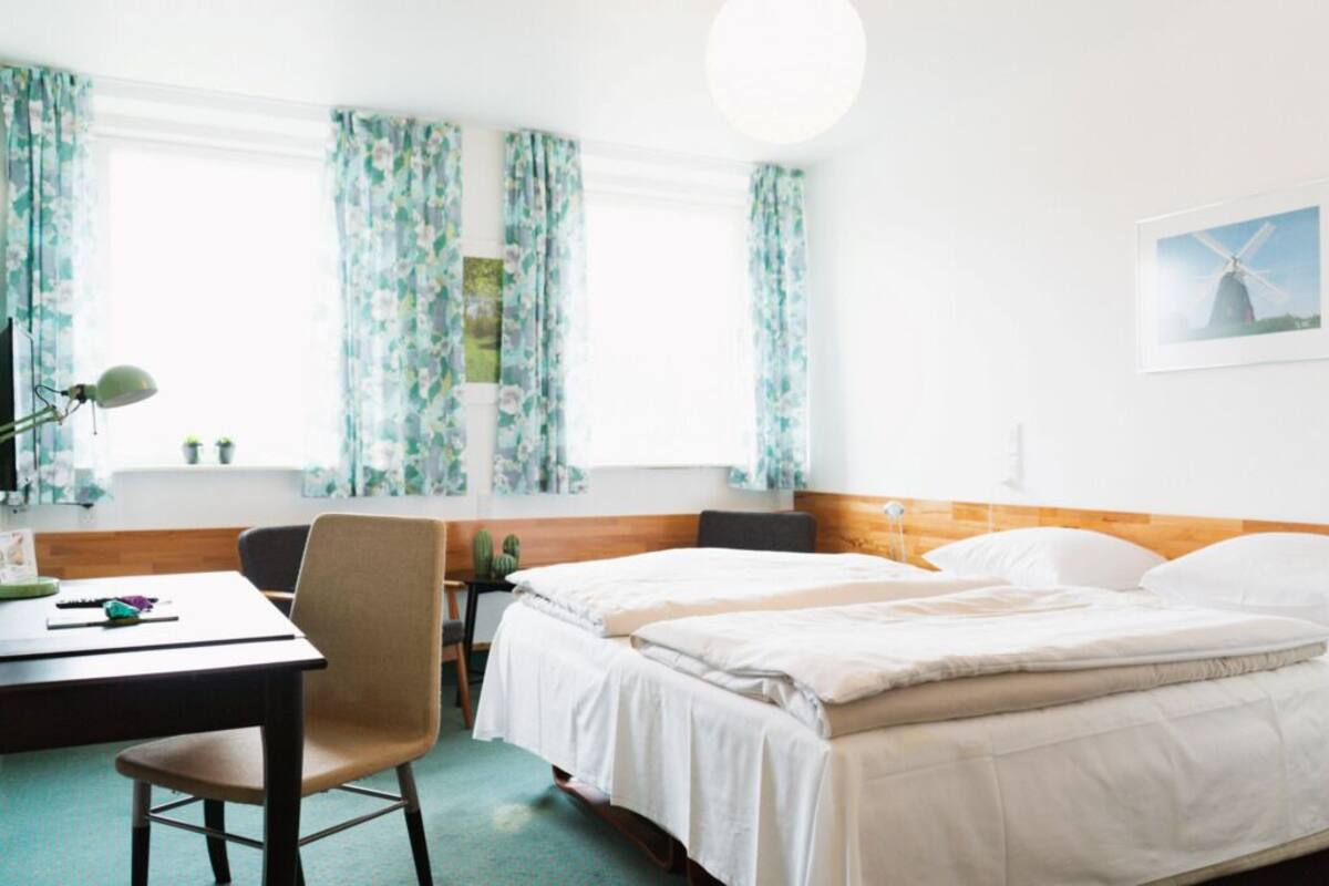Habitación de hotel con dos camas, un escritorio con una silla, una lámpara y un ventanal con cortinas estampadas.